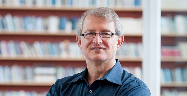 Headshot of Patrick Heller in a blue collared shirt in front of a colorful bookcase.