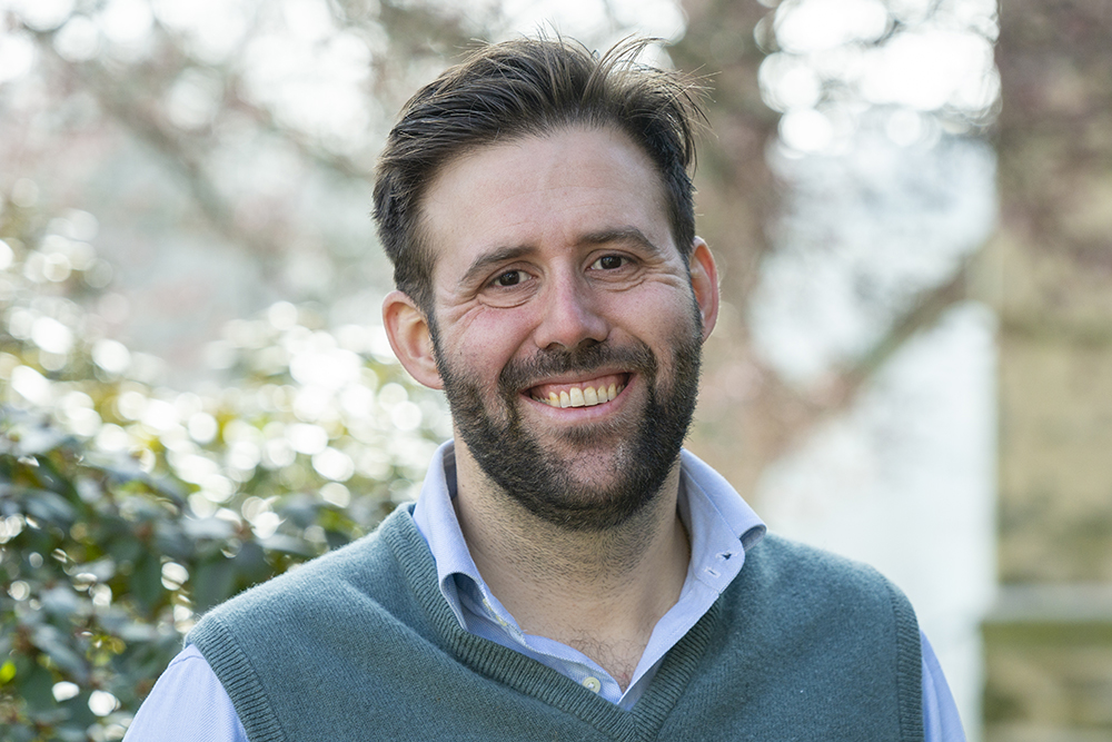 Dennis Hogan in a green sweater vest and blue shirt, smiling with trees in the background