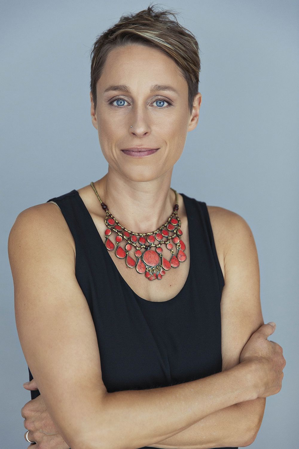 Headshot of Jessica Plavicki wearing a black sleeveless shirt and large orange necklace with a plain blue background