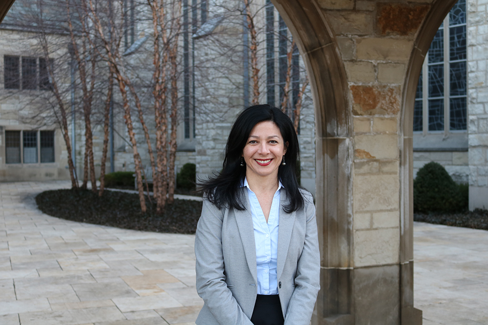 Dr. Lackey sitting by a stone pillar with bare trees in the background; she is wearing a grey suit jacket and blue shirt.
