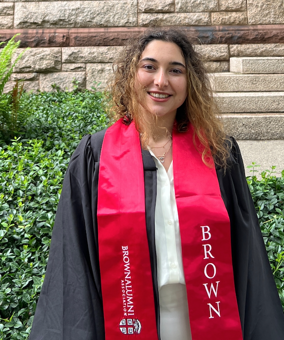 Ilayda Top, smiling in a black graduation gown with a red sash that says Brown Alumni Association and Brown, in front of a brick wall with greenery.