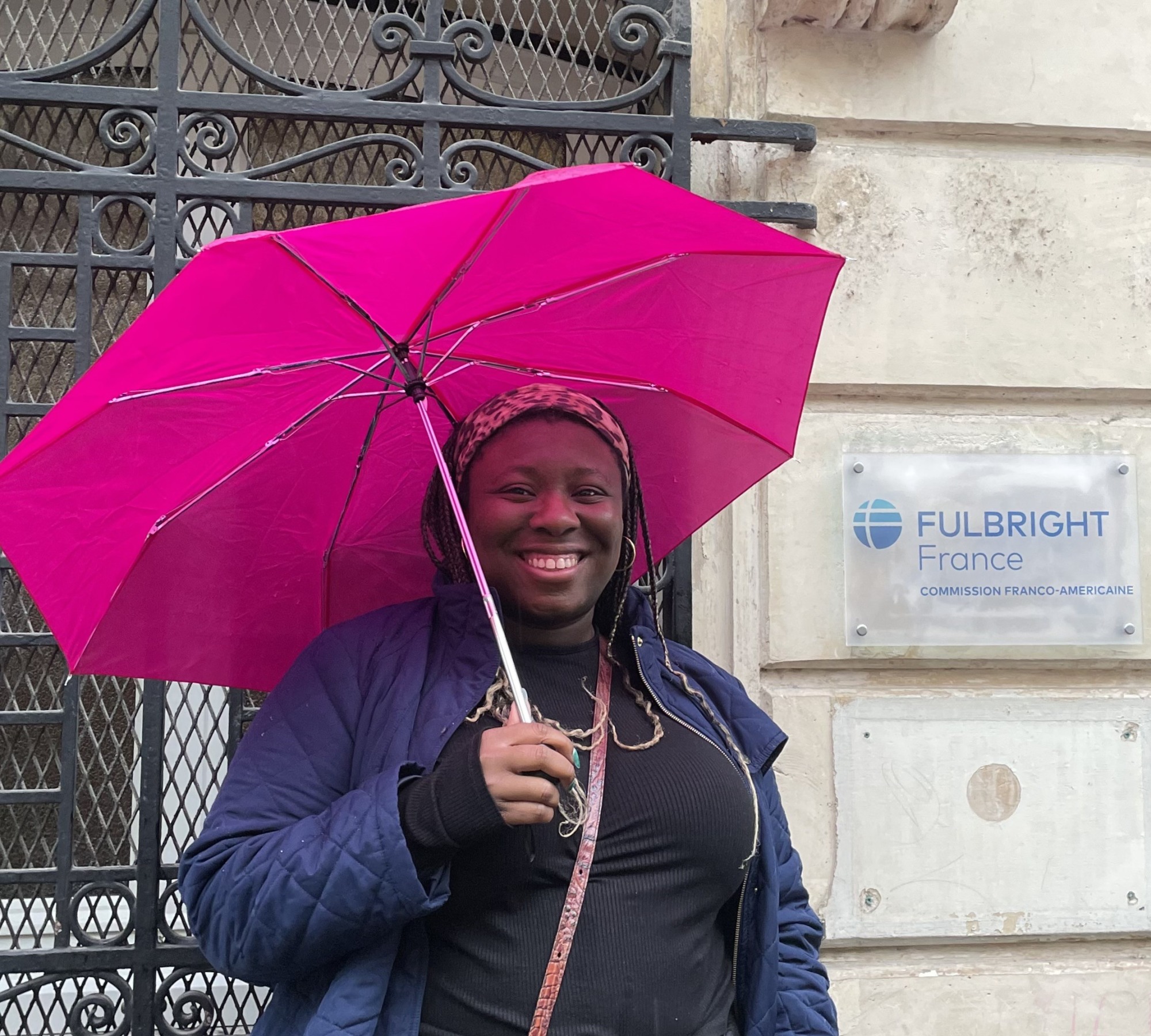 Oluwasemilore Sobande with a dark jacket and pink umbrella outside Fulbright France office