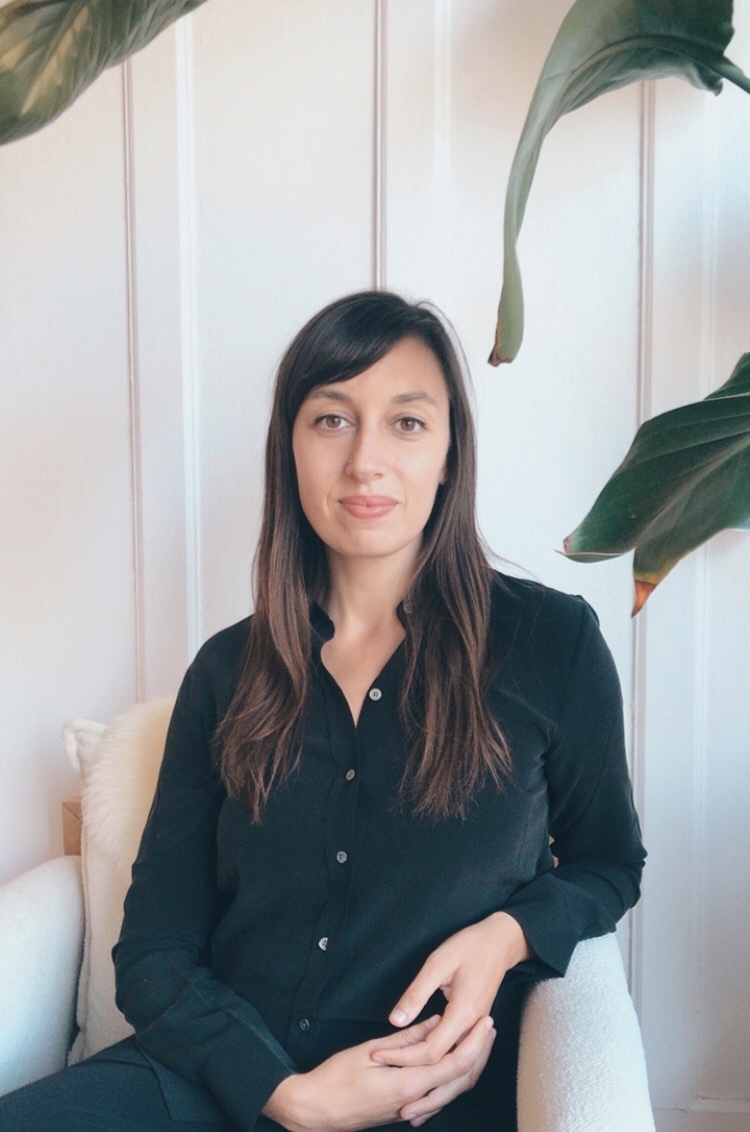 Image of kristen iemma sitting in a chair, with plant leaves beside her; she is wearing a black shirt and pants.