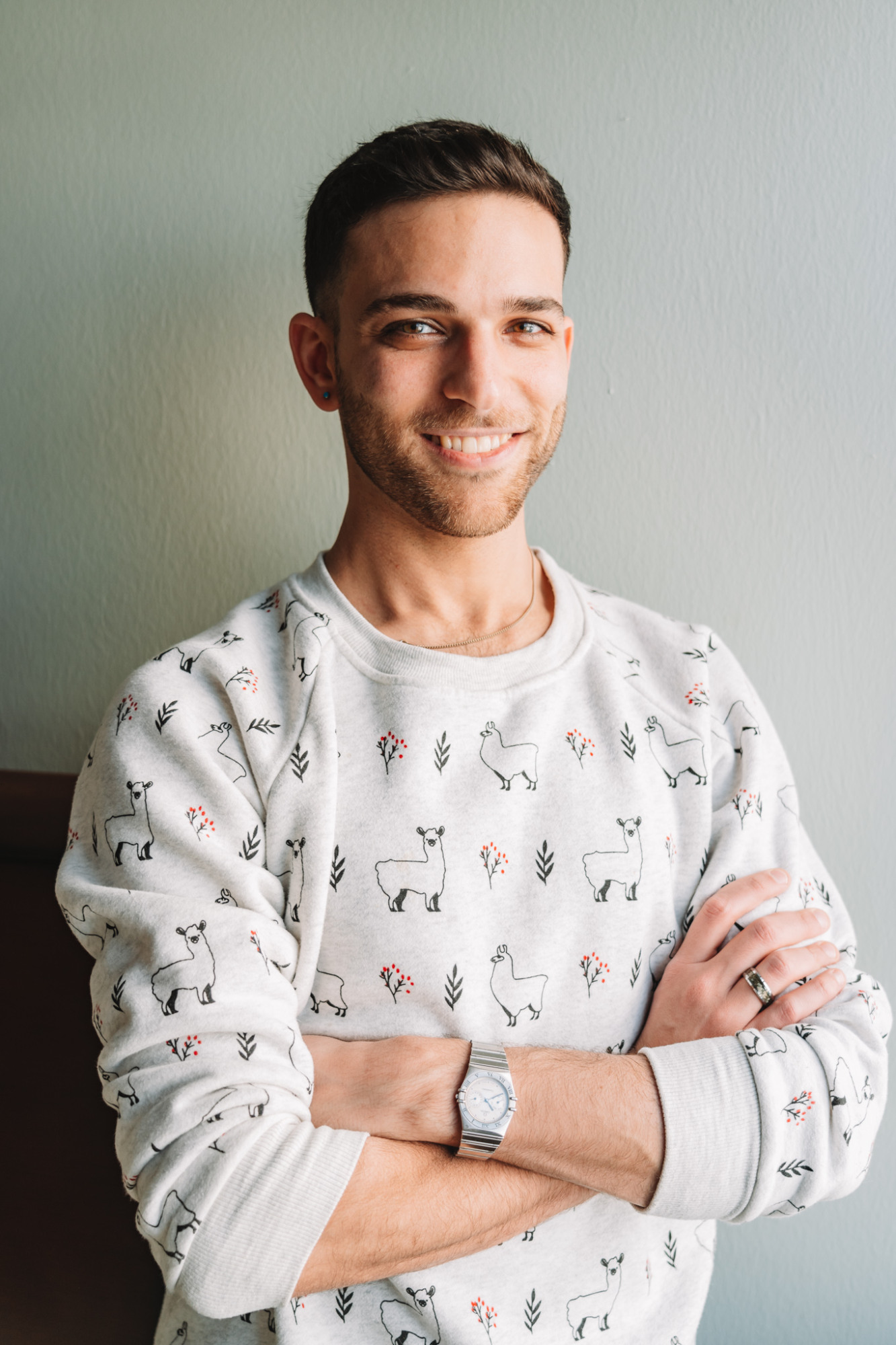 Headshot of Alexander Hardan with short brown hair and a friendly smile, wearing a cream-colored sweater with a small, repeated pattern, posing with his arms crossed.