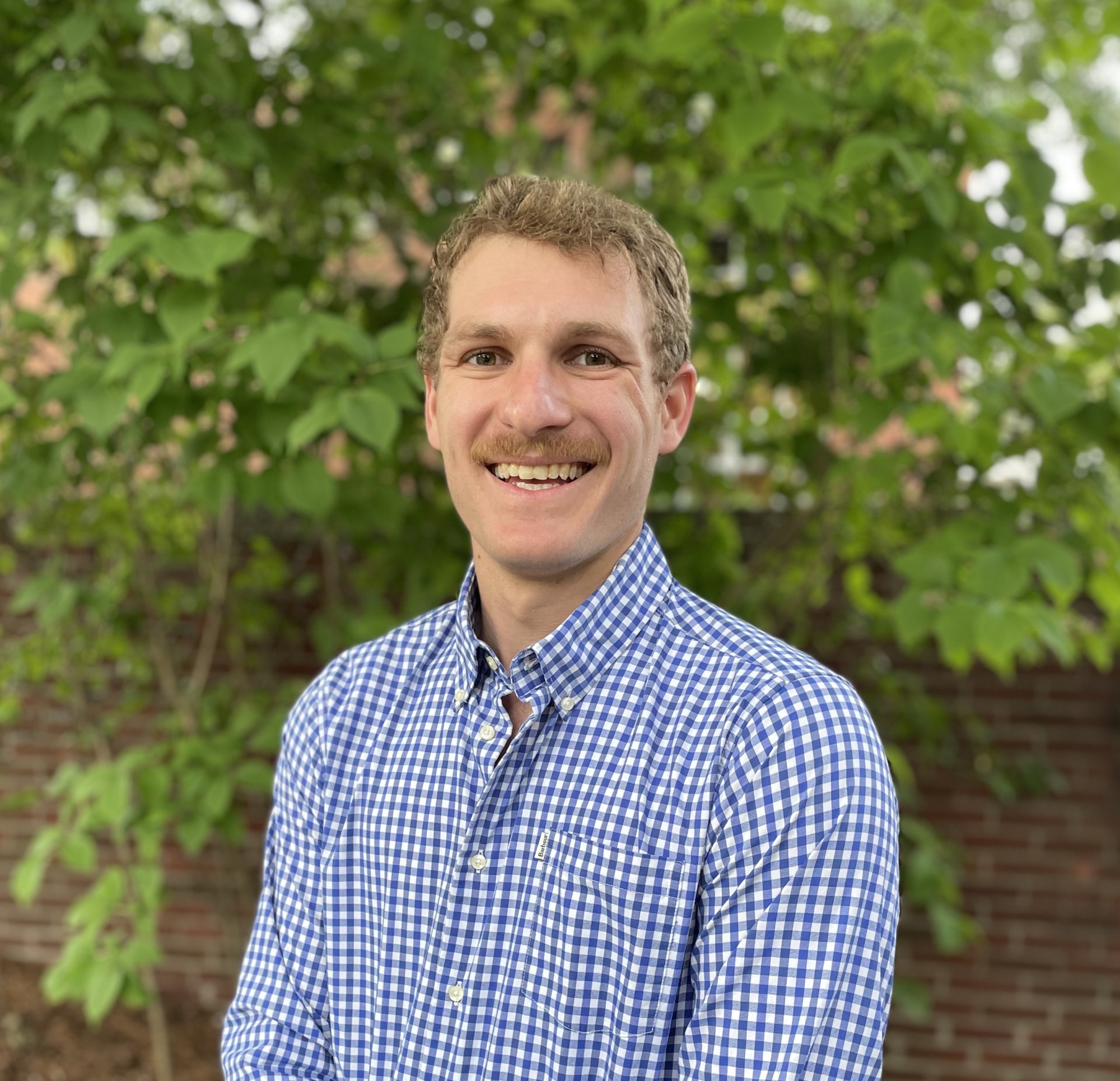 Matthew Ballance with short, wavy brown hair, wearing a blue and white gingham button-down shirt, smiles gently at the camera. He is positioned outdoors against a background of green foliage and a red brick wall.