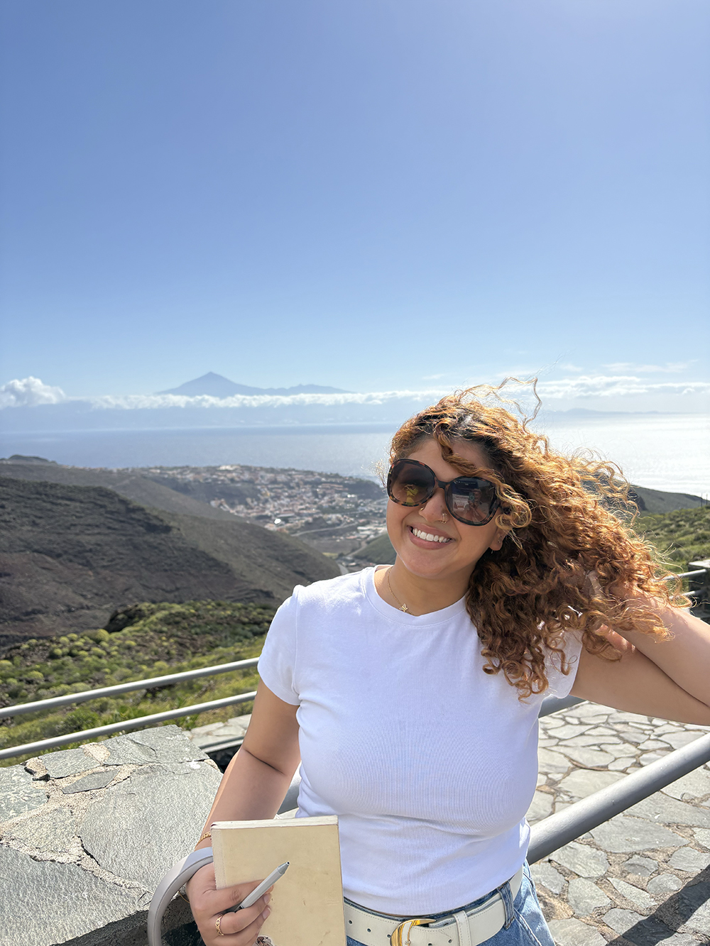 Imen Boussayoud with curly sunlit hair and oversized dark sunglasses stands outdoors on an elevated stone scenic overlook, wearing a white short-sleeved t-shirt and light blue jeans with a white belt. She holds a beige notebook and pen in one hand while running her other hand through her hair, with a vast blue ocean, distant mountains, and a clear sky forming the backdrop behind her.