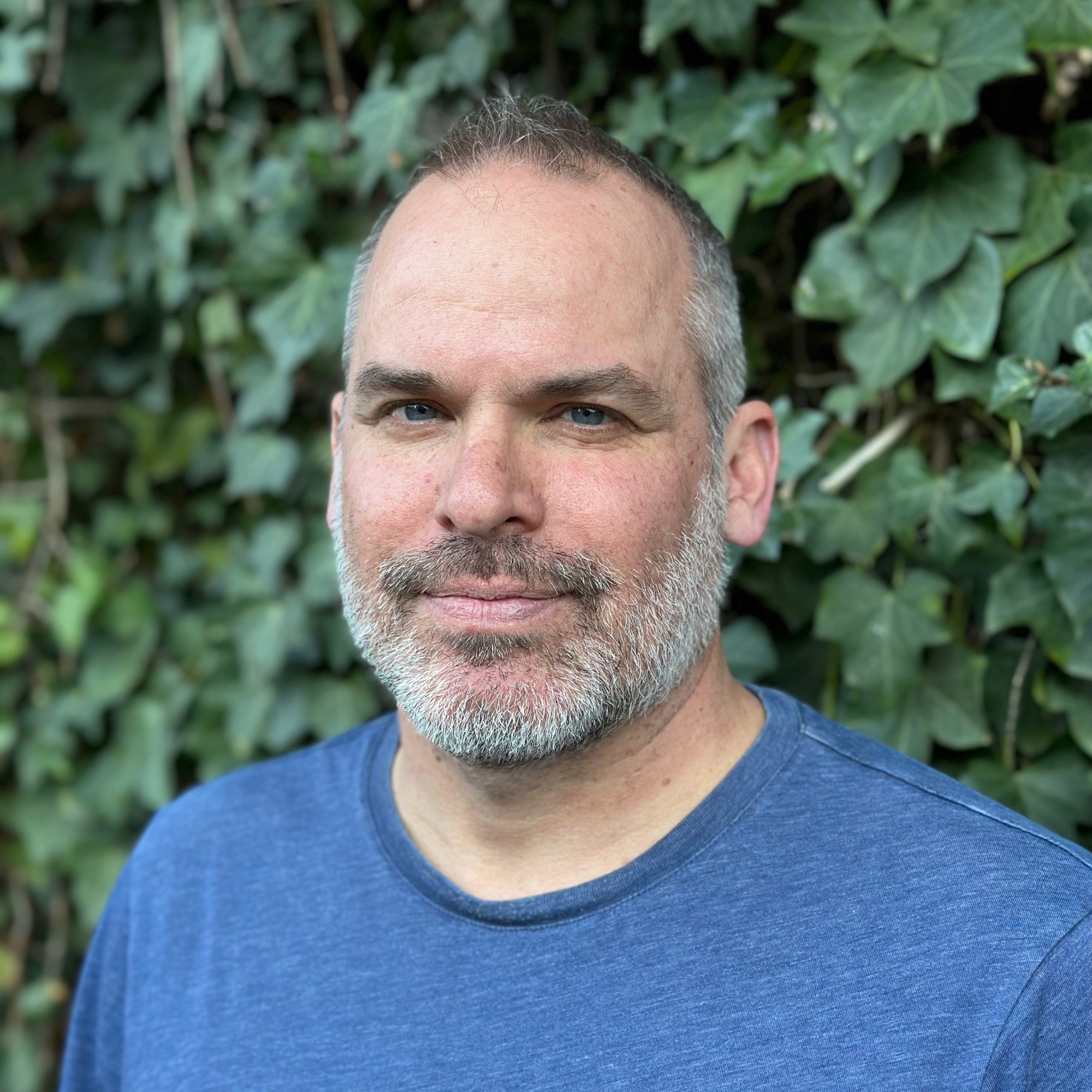 Stephen Bush, Professor and Chair of Religious Studies, smiling and wearing a blue shirt, standing outdoors in front of green ivy.