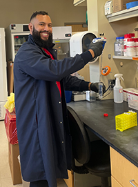 Kelvin in a dark lab coat, smiling with lab equipment in the background