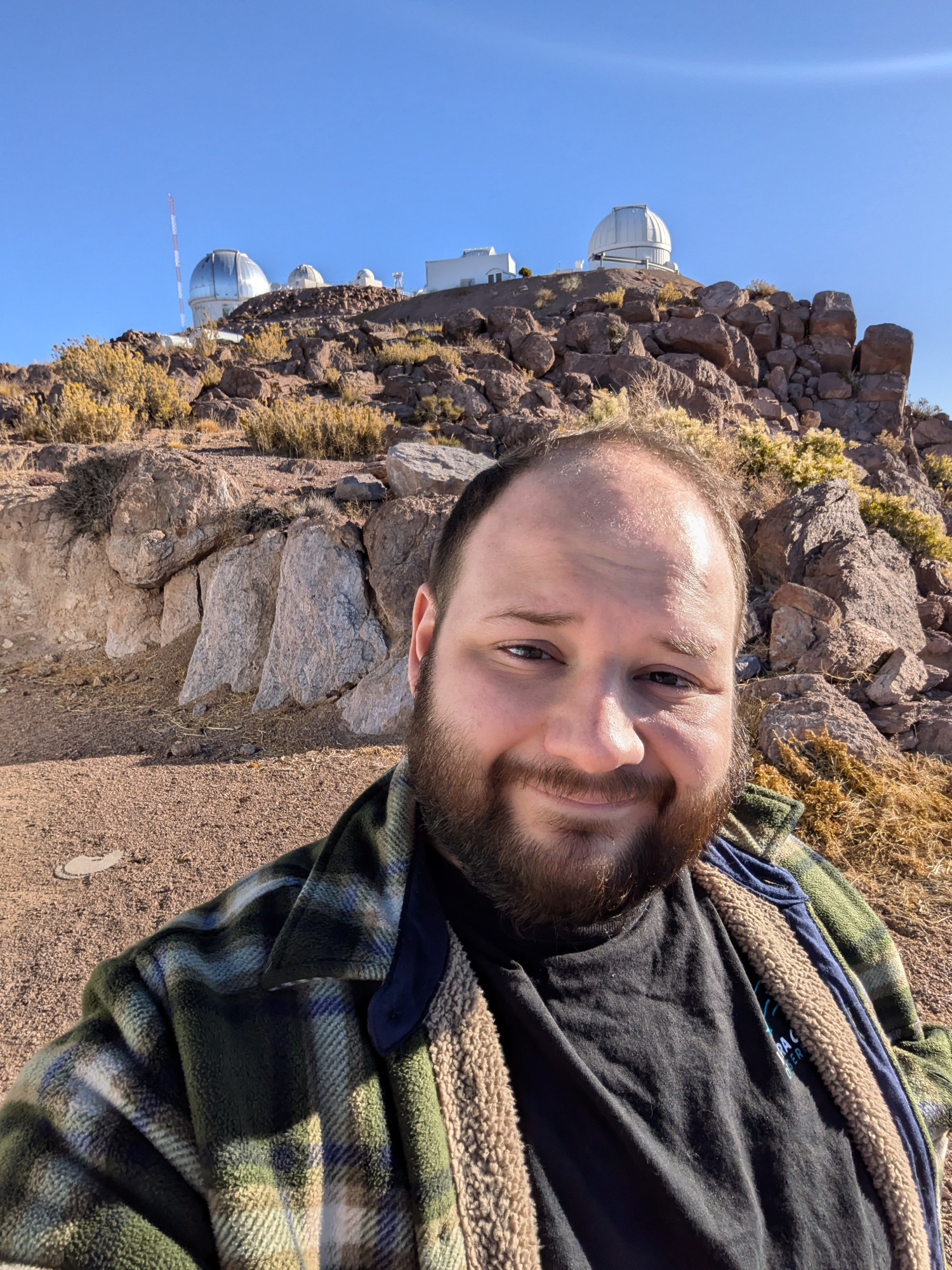 Anthony Englert, wearing a dark jacket, standing outside in a mountainous, rocky landscape with an astronomical observatory dome in the background.