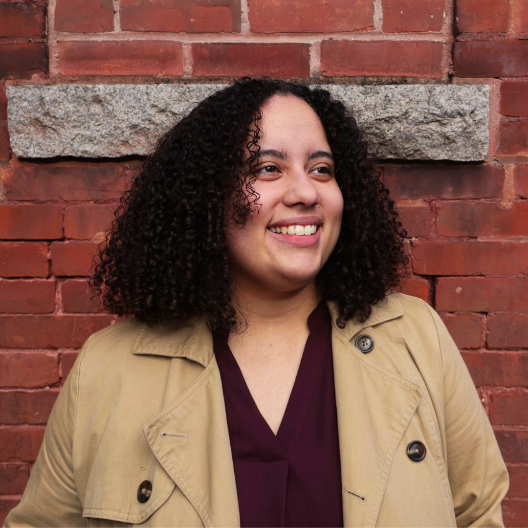Melanie Ortiz Alvarez de la Campa with dark curly hair smiling and looking away, wearing a tan trench coat over a plum top.