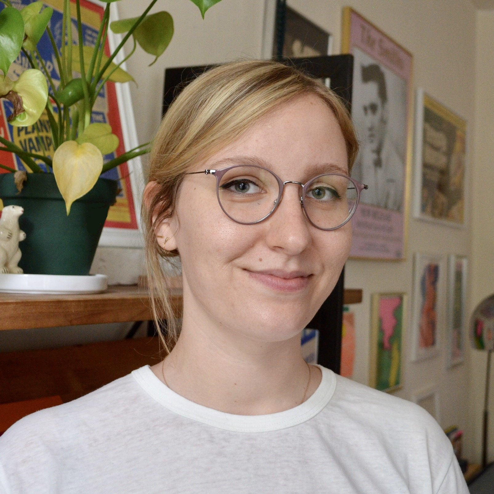 Emily Hirsch with blonde hair tied back and side-swept bangs smiles gently at the camera. She wears round, thin, light grey-tinted wire glasses and a simple white crewneck t-shirt. To her left, a leafy green potted houseplant with a yellowing spade-shaped leaf sits on a wooden shelf, alongside framed posters and artwork decorating the wall in the background.