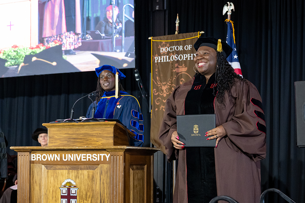 A graduate stands smiling beside a podium holding a black case with the Brown logo, while a presenter in academic regalia addresses the audience from a Brown University podium.