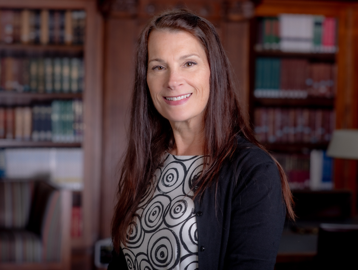 A professional headshot of Michele Welindt smiling, featuring long dark hair and wearing a dark blazer over a patterned top, set against a background of bookshelves.