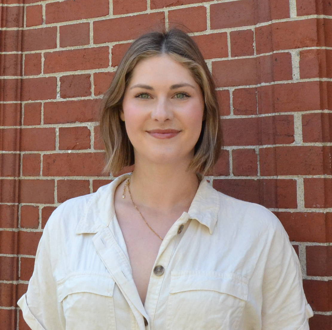 Lulu Schulz, a Ph.D. candidate, smiling and looking at the camera, wearing a light-colored button-down shirt in front of a brick wall.
