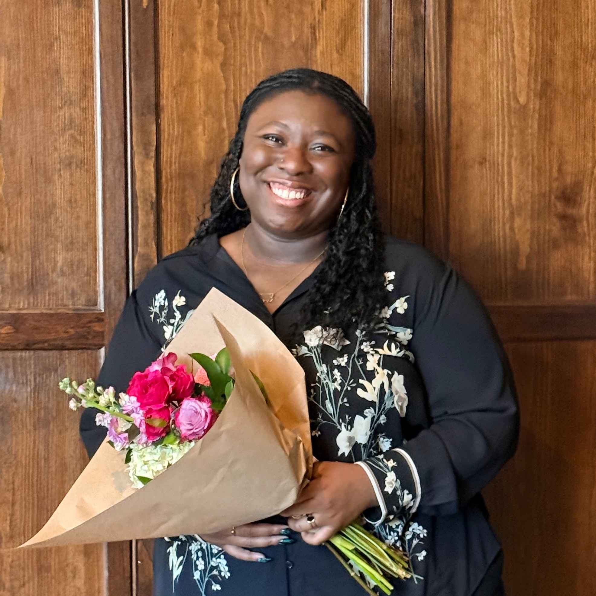 A portrait of Semilore Sobande smiling and holding a large bouquet of colorful flowers. She is wearing a black floral-patterned top and stands in front of a neutral, indoor background.