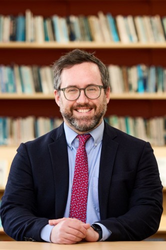 Paul Testa headshot in a dark suit jacket and red tie in front of a bookshelf