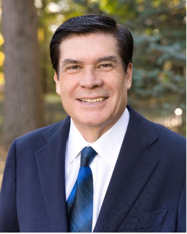 A professional headshot of Horacio Espinosa, a man with dark hair, wearing a dark suit, white dress shirt, and a blue patterned tie, smiling against a light blue background.