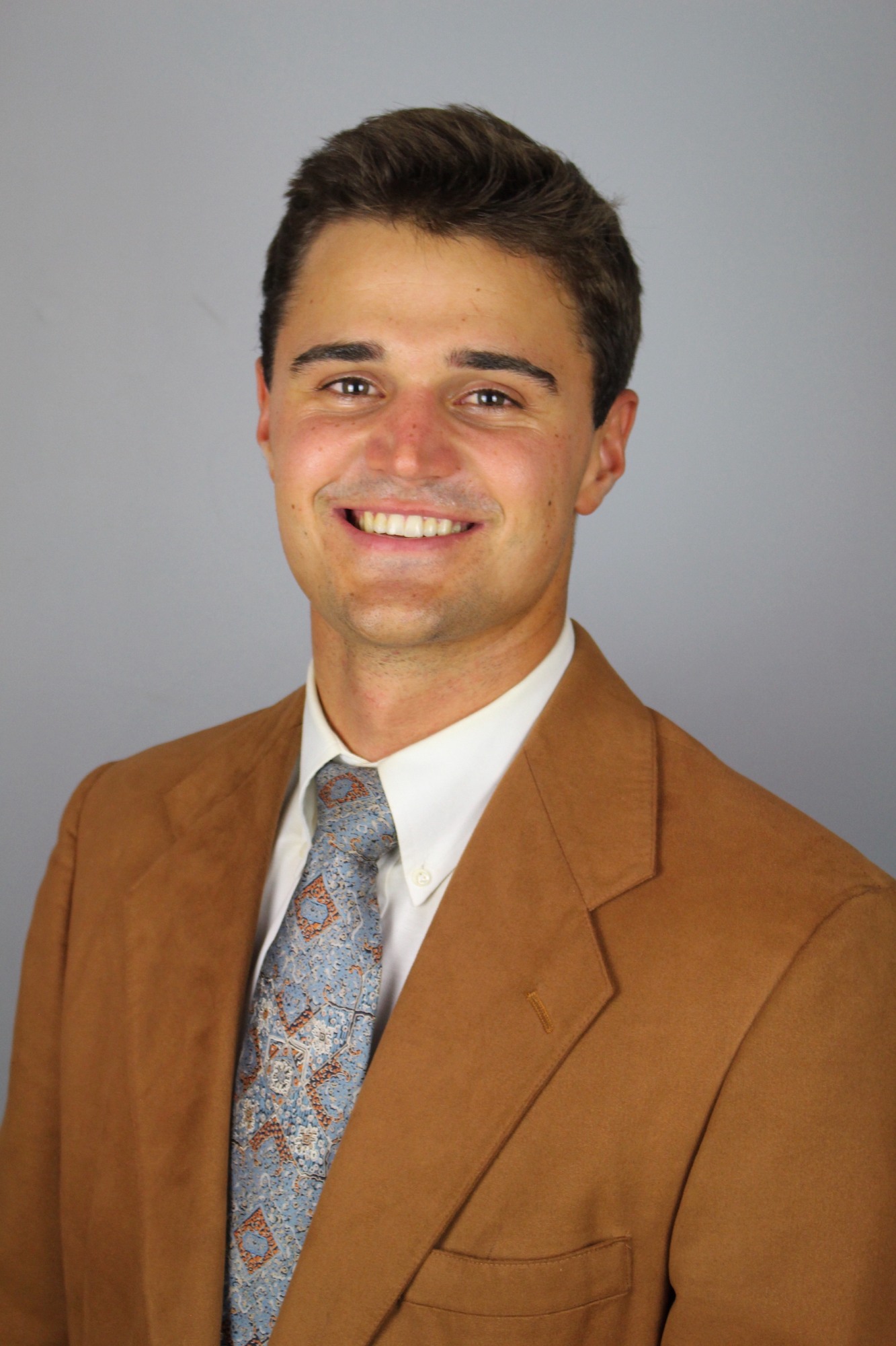 Owen Manahan headshot; Owen is wearing a white shirt, tie and brown jacket with a plain background