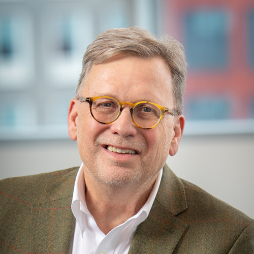 Headshot of Dean Lindstrom with short hair and round tortoiseshell glasses, wearing a white shirt and a green tweed blazer with faint brick windowpanes in the background.