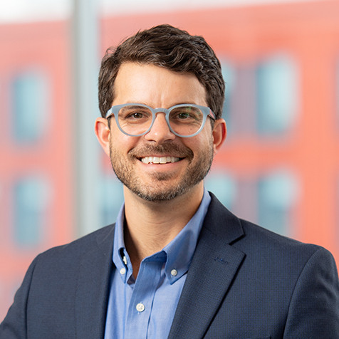 Headshot of Byrd McDaniel with short dark hair, glasses, and a beard stands in a professional setting wearing a blue button-down shirt and a navy blazer. The background features a large window with a blurred view of a modern orange and white building.