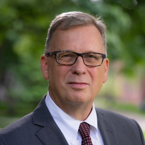 Headshot of David P. Lindstrom wearing glasses, a dark suit jacket, a white shirt, and a dark red tie, with a soft-focus background of green foliage.