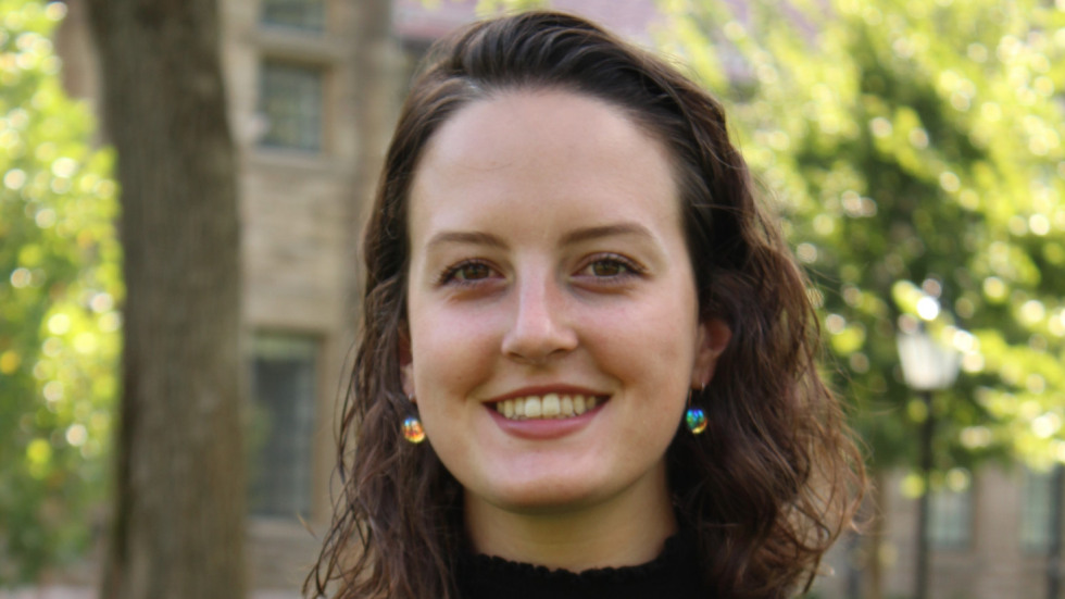 Headshot of a Riley Havel with shoulder-length wavy dark hair, wearing a black shirt and gold drop earrings, standing outdoors in front of trees and a stone building.