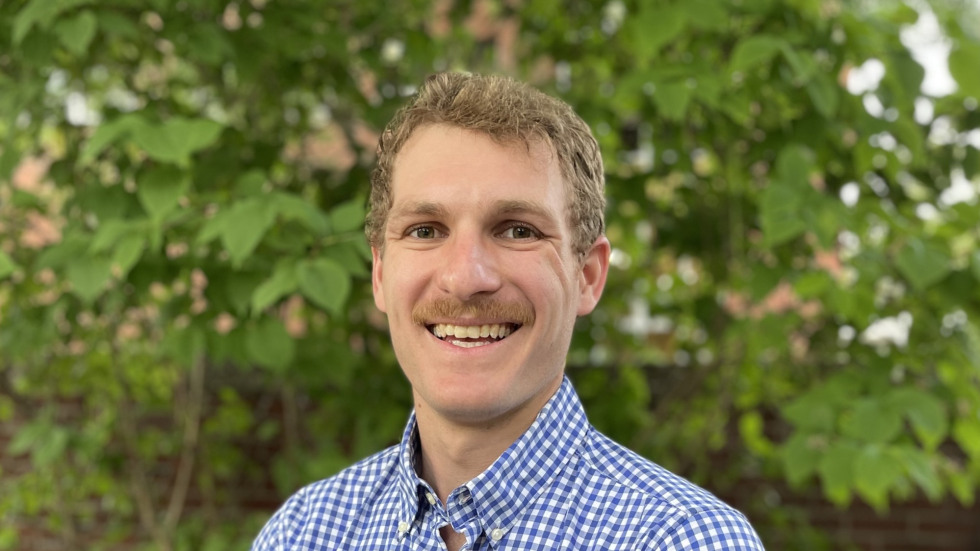 Matthew Ballance with short, wavy brown hair, wearing a blue and white gingham button-down shirt, smiles gently at the camera. He is positioned outdoors against a background of green foliage and a red brick wall.