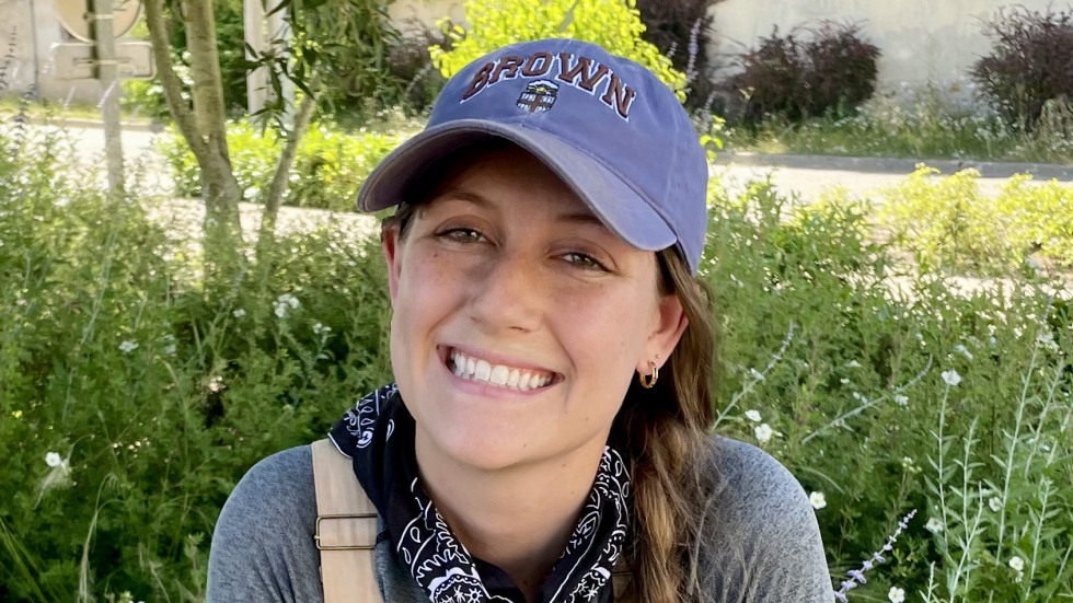 Alyssa Bolster in a gray rain jacket and red pants, wearing black rubber boots and a baseball cap, stands on a mossy, uneven stone step in a lush, overgrown, mountainous landscape. She is partially framed by a dark, stone, cave-like entrance or ruin, which is surrounded by thick green and yellow vegetation.