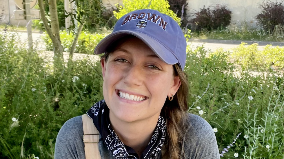 Alyssa Bolster in a gray rain jacket and red pants, wearing black rubber boots and a baseball cap, stands on a mossy, uneven stone step in a lush, overgrown, mountainous landscape. She is partially framed by a dark, stone, cave-like entrance or ruin, which is surrounded by thick green and yellow vegetation.